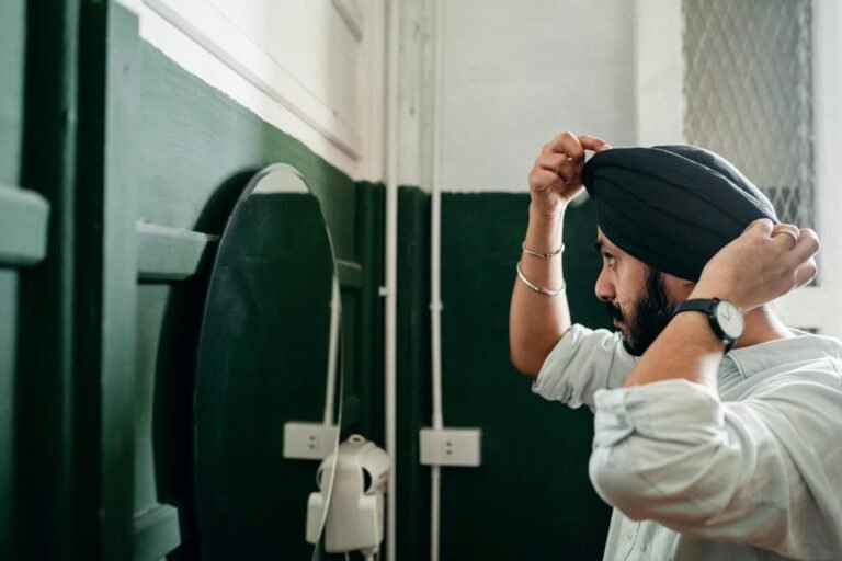 Side view of confident bearded Indian male wearing denim shirt adjusting traditional headwear standing in front of mirror on wall at home while taking care of appearance